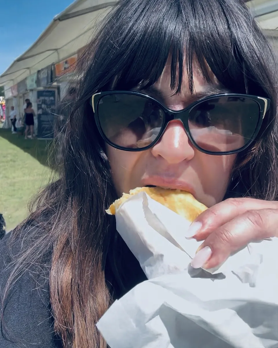 Laura in sunglasses biting into a pasty from a paper bag, a row of market stalls behind her