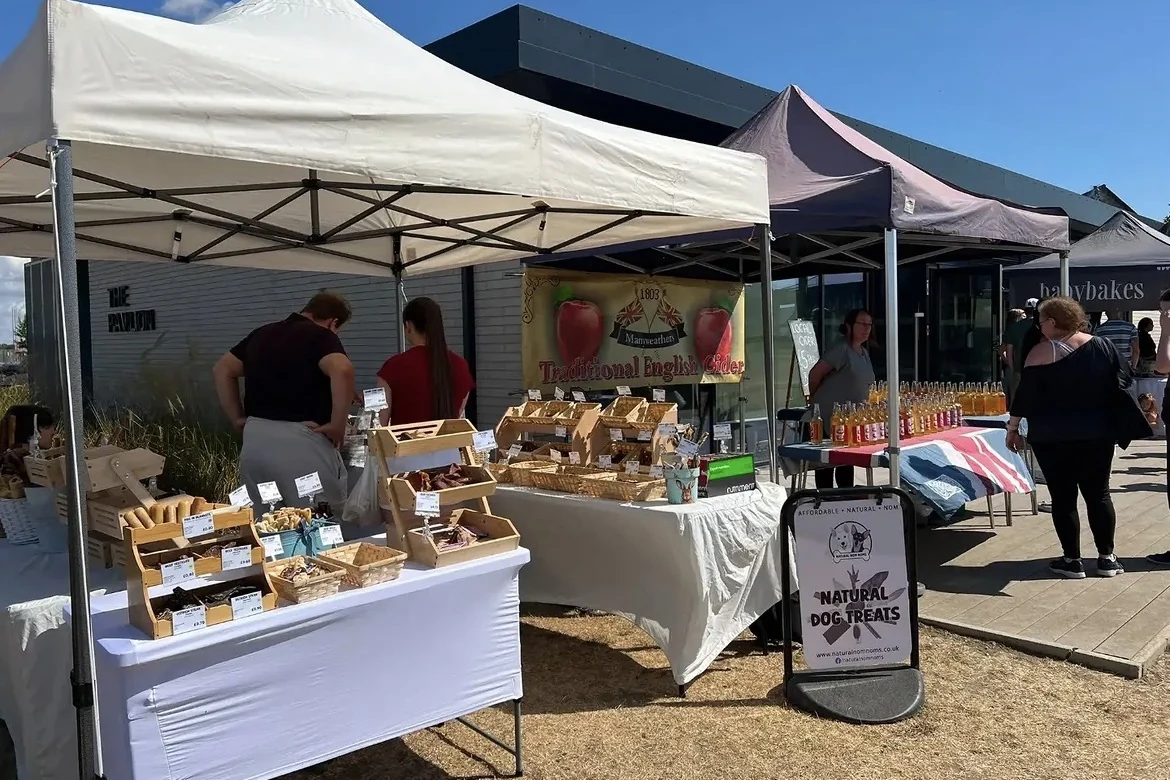 Market gazebos in the sunshine outside The Pavilion: a dog treats stall, traditional English cider in bottles, and shoppers browsing
