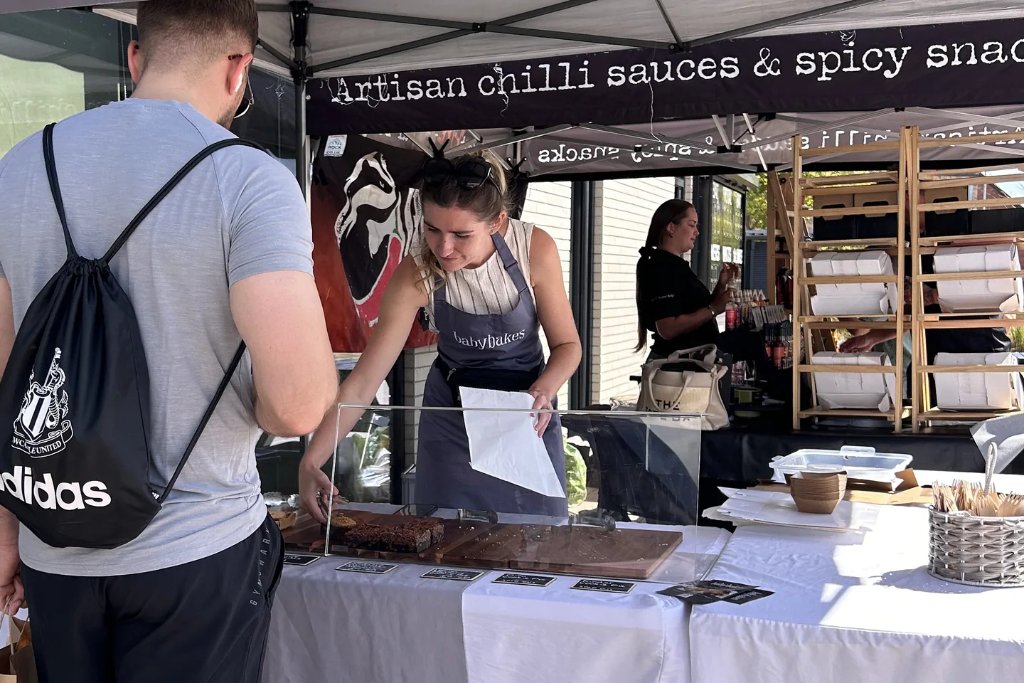 The babybakes trader in her apron serving a customer a brownie from under her stall’s chilli sauces banner