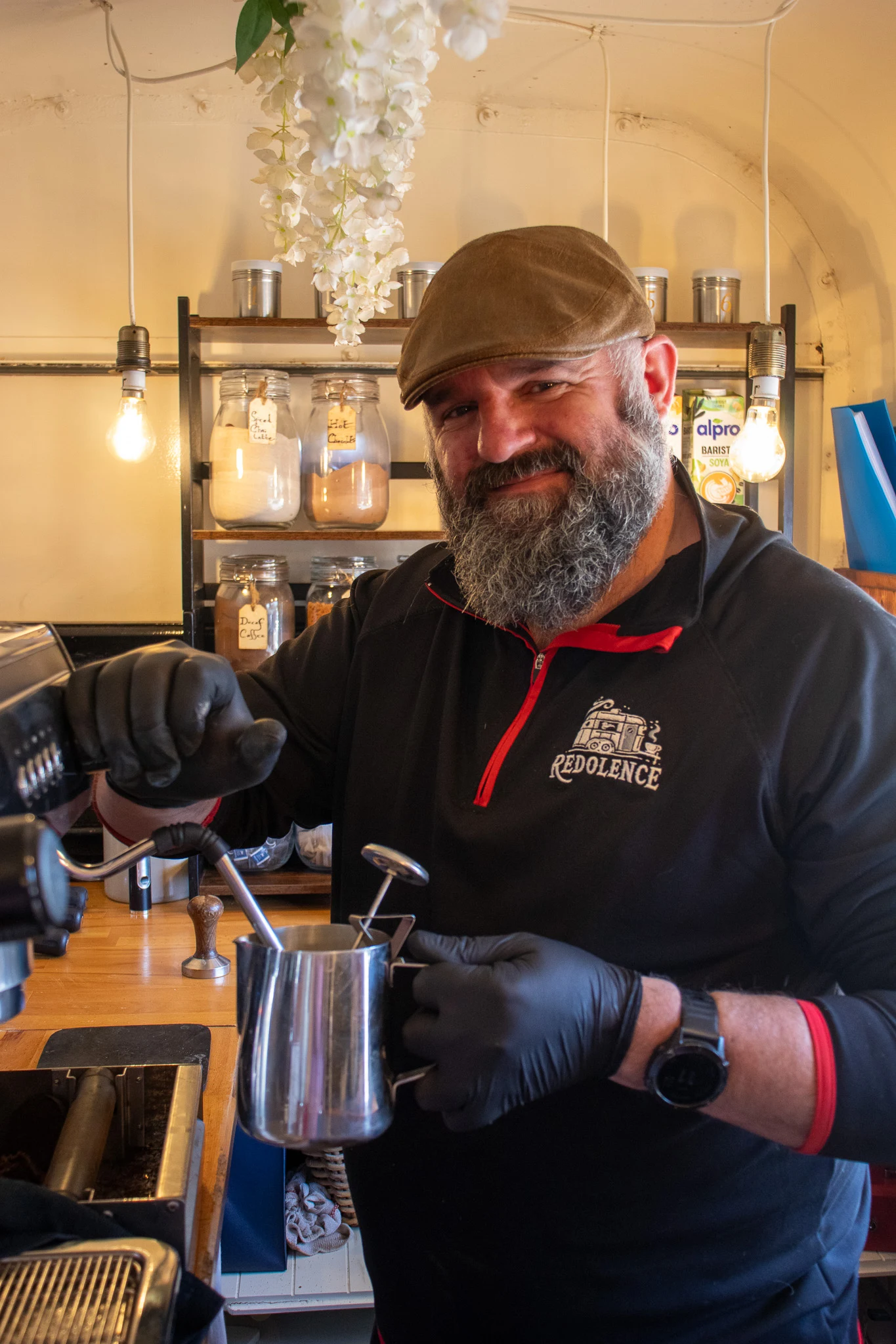 A smiling coffee trader holding a milk jug beside his espresso machine