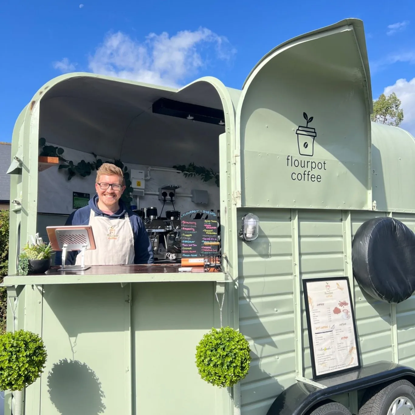A smiling trader serving from a sage-green coffee trailer under a blue sky